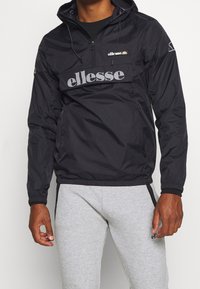 Mann trägt eine schwarze ellesse-Windjacke mit Frontreißverschluss und großem Logo, kombiniert mit hellgrauen Jogginghosen mit Reißverschlusstaschen.
