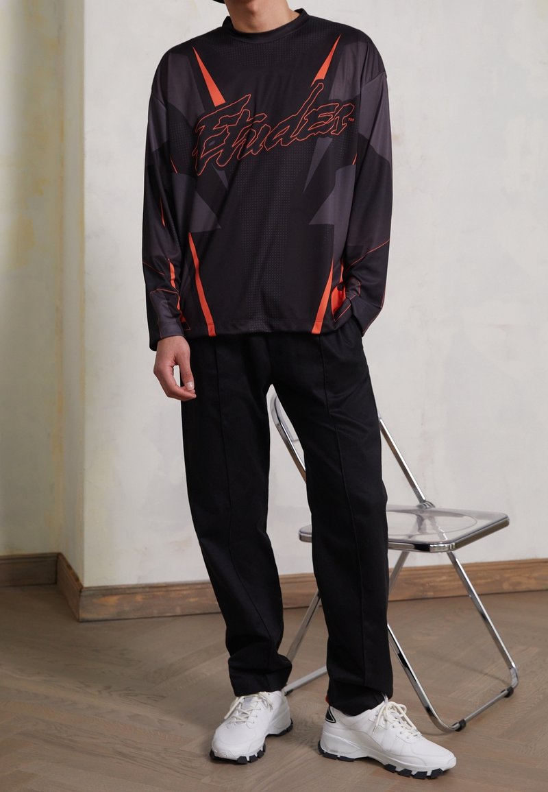 Svart idrottströja med röda detaljer och grafisk text, kombinerad med svarta byxor och vita sneakers. Slät tygkvalitet med en avslappnad passform.