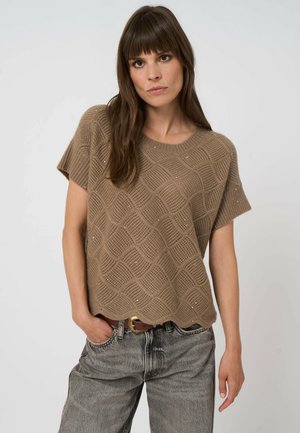 Femme aux longs cheveux bruns portant un pull court à manches courtes en maille texturée de couleur marron clair et un jean gris délavé avec une ceinture marron, une main dans la poche.