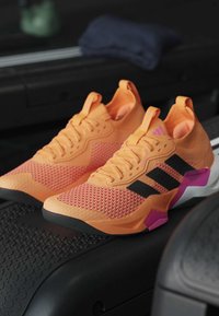 Paire de chaussures de sport orange et rose avec des rayures noires, placées sur une surface noire de tapis roulant.