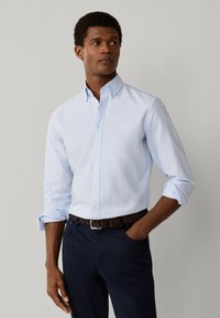 Uomo in piedi con la mano in tasca, che indossa una camicia azzurra a bottoni e pantaloni scuri su uno sfondo grigio semplice.