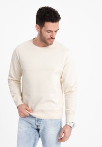 Felpa crema con maniche lunghe, polsini e scollatura a costine. Realizzata in morbido tessuto, presenta una vestibilità comoda. Indossata con jeans azzurri.