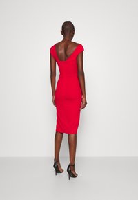 Kobieta w dopasowanej czerwonej sukience midi z szerokim dekoltem typu off-shoulder i czarnych sandałach na wysokim obcasie, stojąca na jednolitym tle.