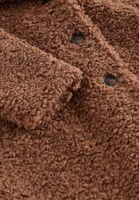 Manteau en polaire marron texturé avec une surface duveteuse, de grands boutons et des plis superposés au niveau du col et de l'avant.