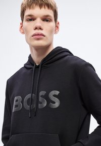 Μαύρο αθλητικό φούτερ με κουκούλα και μπροστινή τσέπη καγκουρό. Διαθέτει μεγάλο, γυαλιστερό λογότυπο "BOSS". Το υλικό φαίνεται μαλακό και λείο, με κουκούλα που δένει με κορδόνια.
