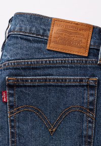 Pantaloni in denim blu con tasca posteriore foderata di cuciture arancioni, etichetta rossa Levi's e marchio in pelle marrone della Levi Strauss & Co. sopra la tasca.