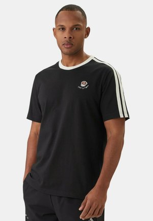 Hombre vistiendo camiseta negra con cuello blanco y rayas en las mangas, con las manos en los bolsillos, de pie frente a un fondo liso.