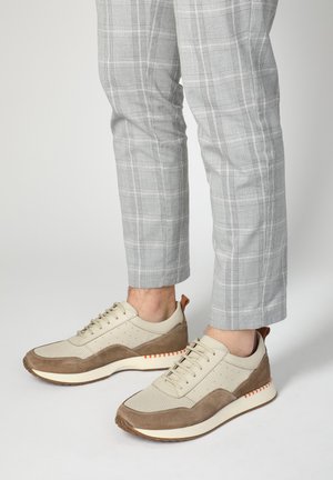 Beige- und braune Sneaker mit einem strukturierten Mesh-Obermaterial, Lederelementen, Perforationen für Atmungsaktivität und einem kontrastierenden orangefarbenen Streifen an der Sohle.