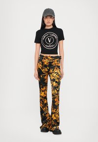 Camiseta corta negra con un logo central, combinada con pantalones de terciopelo acampanados negros y dorados con estampado y zapatos de plataforma negros. Lleva una gorra gris.