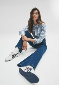 Gestreiftes Hemd in Blau und Weiß mit Knopfverschluss, kombiniert mit dunklen Schlagjeans. Weiße Sneaker mit roten und blauen Akzenten.