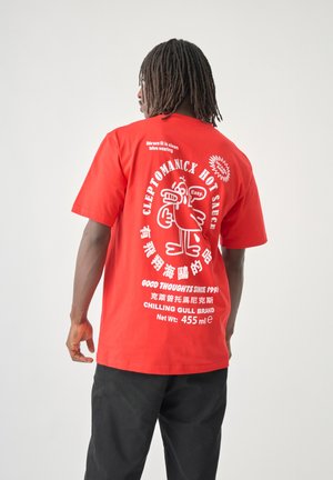 Person with dreadlocks wearing a red T-shirt with white hot sauce-themed graphic text and cartoon bird on the back, paired with black pants.