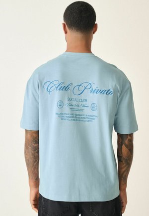 Himmelblå bomulds T-shirt med korte ærmer og rund hals. Har en stor trykt tekst på ryggen i en mørkere blå nuance samt yderligere detaljer nedenfor.