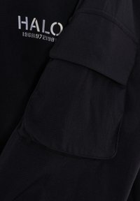 Schwarzes Langarmshirt mit einer aufgesetzten Tasche am Ärmel. Auf der Vorderseite steht in moderner Schrift der weiße Text "HALO 196819721988". Strukturiertes Gewebe.