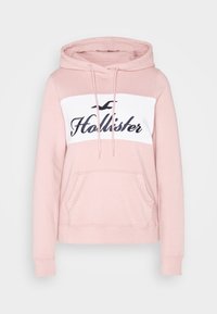 Světle růžová mikina s klokaní kapsou vpředu, šňůrkami na kapuci, bílou částí přes hrudník s černým logem Hollister a ikonou racka.
