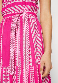 Robe rose avec des motifs géométriques blancs, dotée d'une ceinture à la taille et d'une jupe plissée. Le tissu est léger et texturé.