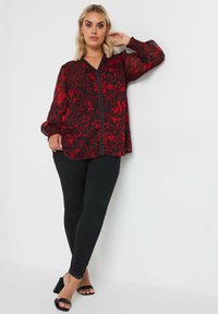 Schwarze transparente Bluse mit rotem Blumenmuster, V-Ausschnitt und Knopfdetails an der Vorderseite. Kombiniert mit schwarzen eleganten Hosen und Sandalen mit Absätzen.