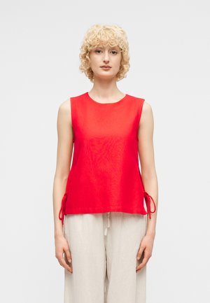 Femme aux cheveux blonds courts et bouclés, portant un haut rouge sans manches avec des liens sur les côtés et un pantalon beige ample, debout devant un fond uni.