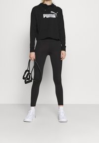 Svart croppad hoodie med vit "PUMA"-logga, matchad med svarta leggings och vita sneakers. Modellen håller en svart träningsaccessoar.