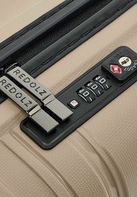 Beige harde koffer met een zwart combinatieslot, twee zilveren ritsen met het label "REDOLZ" en een getextureerd oppervlak.