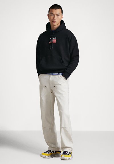 Svart hettegenser med amerikansk flaggmotiv, beige vide bukser, og fargerike joggesko med mønsterdetaljer. Avslappet streetwear-stil.