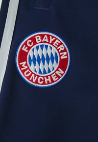Το κεντημένο λογότυπο της FC Bayern München με μπλε και άσπρο διαμαντένιο σχέδιο σε σκούρο μπλε ύφασμα και λευκή ρίγα.