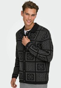 Svart mönstrad cardigan med en texturerad stickad design med geometriska former och krage. Knappar fram och ribbade ärmslut.