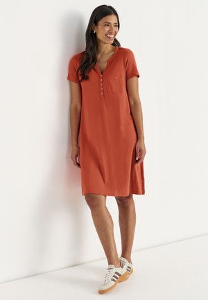 Kvinde smilende, stående, iført en kortærmet, brændt orange knælængde kjole med knapper og en brystlomme, hvide sneakers med striber.