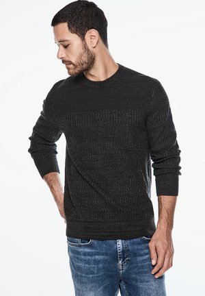 Donkergroene crewneck trui met een gestructureerd patroon, geribbelde boorden en zoom, gecombineerd met lichtblauwe jeans. Model staat met de hand op de heup.