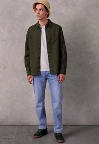 Cămașă button-up de culoare verde măslinie peste un tricou gri, blugi ușor albaștri și pantofi verzi închisi. Pălăria este bej. Ținută casual cu un croi lejer.