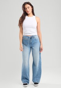 Top corto sin mangas blanco combinado con jeans de pierna ancha en azul claro que cuentan con cordón y detalle de bolsillo frontal. Fondo liso.