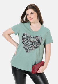 Lysegrøn T-shirt med et stort sort hjerte design, der har paisley mønstre. Korte ærmer, rund halsudskæring, afslappet pasform, parret med en rød clutch.