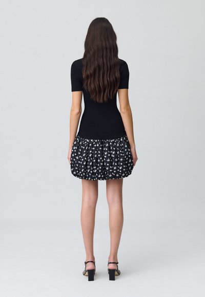 Haut noir ajusté à manches courtes, associé à une jupe noire évasée ornée de motifs floraux blancs. Sandales à talons hauts visibles.