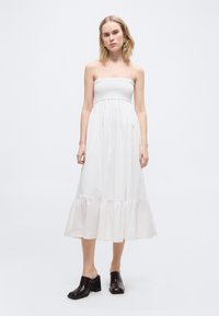 Νεαρή γυναίκα με λευκό φόρεμα μέσης μήκους χωρίς λουριά και με ρεβέρ στο τελείωμα, φοράει μαύρα ψηλοτάκουνα mules και χρυσούς κρίκους σκουλαρίκια, στέκεται μπροστά σε απλό φόντο.