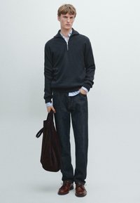 Dunkler Pullover mit halbem Reißverschluss, hellblaues Hemd darunter, dunkle Jeanshose und braune Lederschuhe. Hält eine dunkelbraune Tasche.