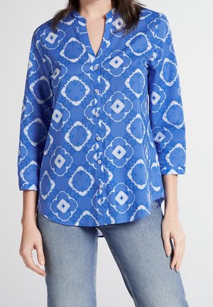 Niebieska koszula zapinana na guziki z białymi geometrycznymi wzorami tie-dye noszona z jasnoniebieskimi dżinsami, przedstawiona od szyi do bioder.