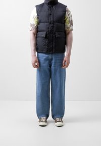 Czarna pikowana kamizelka z wysokim kołnierzem i zapięciem na guziki, noszona na koszulce tie-dye. Luźne, jasnoniebieskie spodnie typu szerokie nogawki oraz brązowe tenisówki.