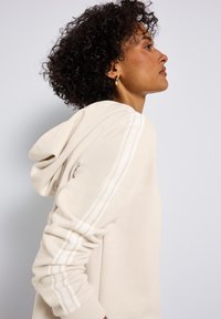 Felpa beige con cappuccio e coulisse, con strisce bianche sulle spalle. Realizzata in morbido tessuto, ha una vestibilità ampia e polsini a coste.