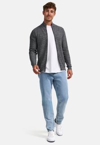 Chaqueta gris con cremallera y cuello, llevada sobre una camiseta blanca. Combinada con pantalones vaqueros azul claro y zapatillas blancas. Tejido suave con un ajuste relajado.