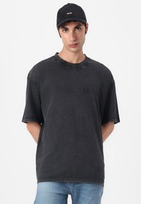 Black oversized T-shirt with distressed texture and a ribbed crew neckline, featuring a subtle logo on the front. Paired with a black cap.