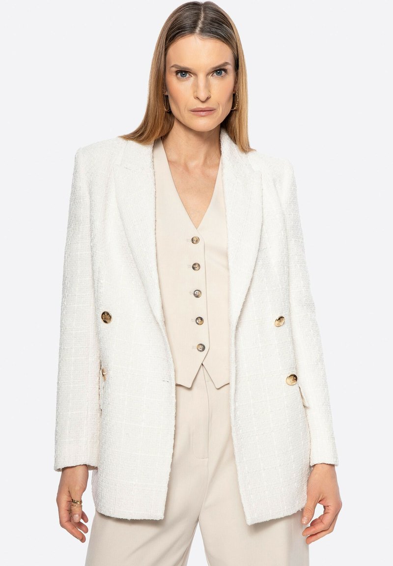 Blazer bianco con texture e vestibilità ampia, dotato di bottoni dorati doppi e un rever strutturato, abbinato a un gilet beige chiaro.