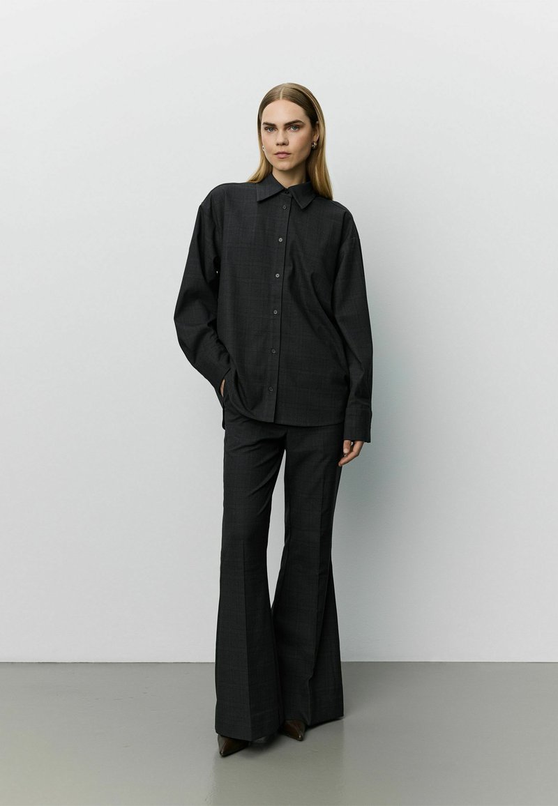 Femme aux cheveux longs et raides, portant une chemise noire ample boutonnée et un pantalon noir à jambes larges, debout contre un mur clair uni.