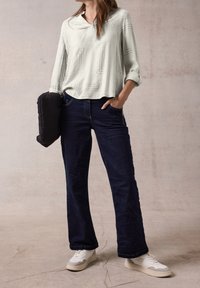 Blouse à manches longues gris clair avec imprimé géométrique, associée à un jean large bleu foncé et des baskets blanches. Tenant un sac noir matelassé.