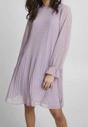 Vrouw die een loszittende, lavendelkleurige jurk draagt met lange mouwen en een lengte tot aan de knieën, met een subtiel patroon en een geplooide taille, voor een effen achtergrond.