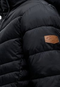 Μαύρο καπιτονέ μπουφάν με οριζόντιες ραφές και ένα μικρό καφέ δερμάτινο ταμπελάκι στο μανίκι.