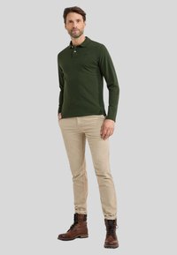 Donkergroene langemouw polo, beige slim fit broek, bruine veterschoenen met zwarte accenten, staand op een neutrale grijze achtergrond.