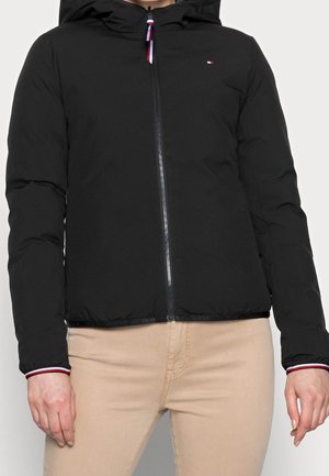 Veste noire à capuche avec fermeture éclair, petit logo sur la poitrine, portée avec un pantalon beige, avec des détails à rayures rouges et blanches sur la tirette de la fermeture éclair et les poignets.