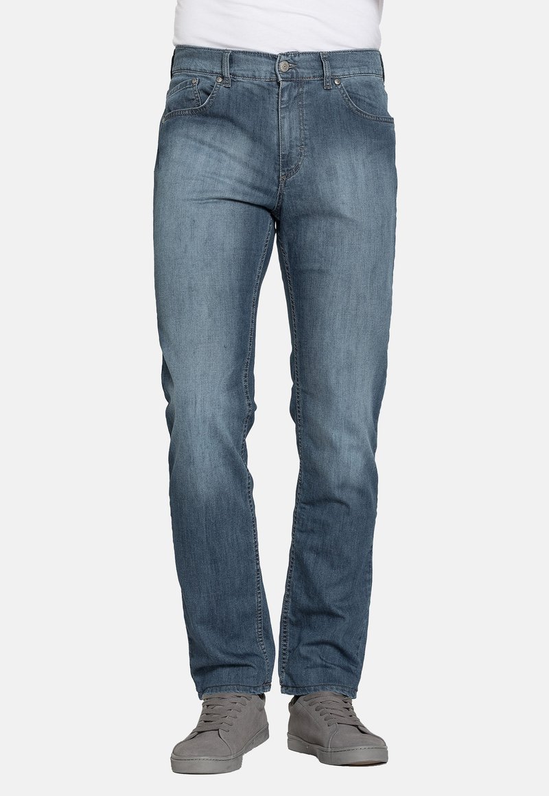 Uomo che indossa jeans blu a gamba dritta e sneaker casual grigie, in piedi davanti a uno sfondo semplice.