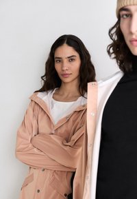 Two individuals are wearing outerwear. One has a light pink waterproof jacket with a hood, and the other wears a black turtleneck.