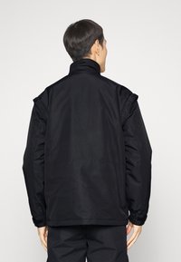 Svart jacka med hög krage, breda axlar och elastiska ärmar. Tyget har en slät yta med en något glänsande finish.