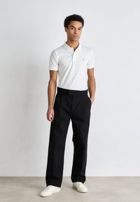 Valkoiset lyhythihaiset polo-paidat, joissa on huomaamaton logo rinnassa, yhdistettynä korkeavyötäröisiin mustiin housuihin ja valkoisiin lenkkareihin.